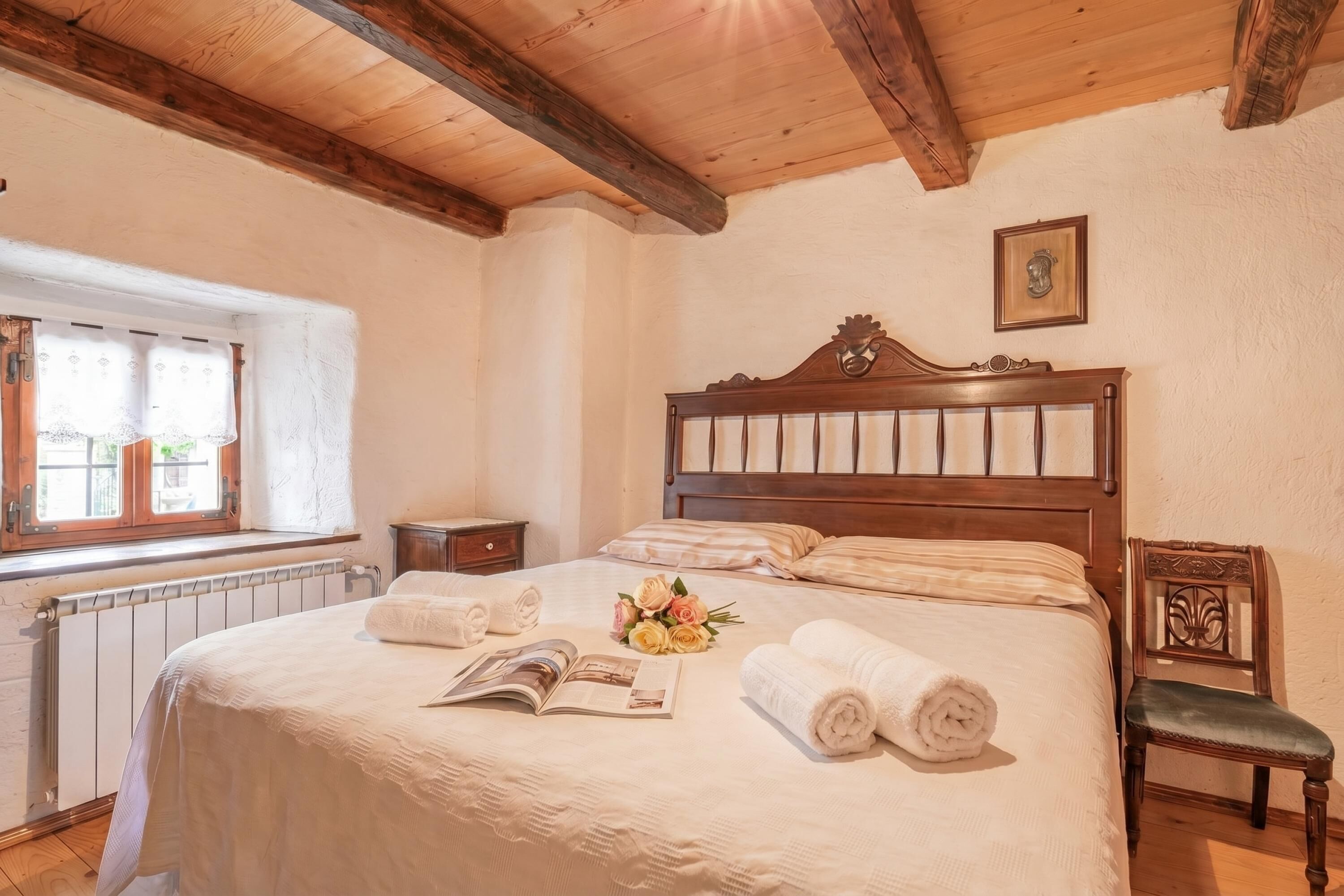 Ein ordentlich bezogenes Bett mit einem Buch und Blumen, ein hölzernes Kopfteil, ein Stuhl und ein Fenster mit Vorhängen.
