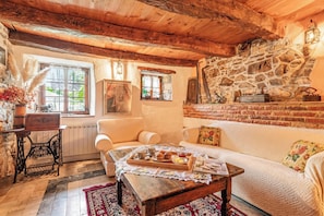 A cozy living room with a stone wall, wooden ceiling beams, a sofa, a coffee table with a tray of pastries, and a vintage sewing machine. - Rustic House Luin (Komen)