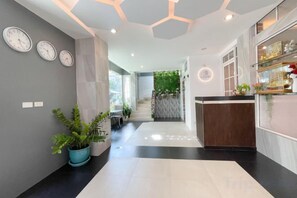 A modern hotel lobby with a central staircase, a reception desk, and a plant on the left. - Le Cartier (Phuket)