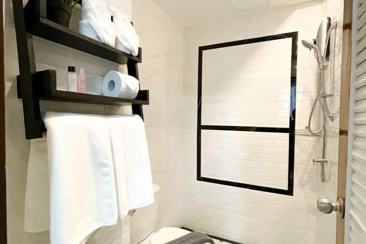Une petite salle de bain propre, avec des carreaux blancs, un porte-serviettes avec des serviettes blanches, une étagère avec des articles de toilette et un espace douche.