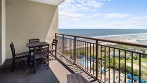 Un balcon avec vue sur l’océan et la plage, des meubles en bois et, au fond, un espace piscine.