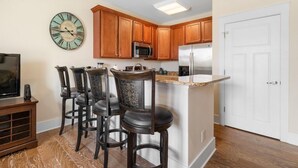 A kitchen with wooden cabinets, a stainless steel refrigerator, a microwave, and a bar stool area. - Carriage Home Charm: A Romantic Getaway for Two (North Myrtle Beach)