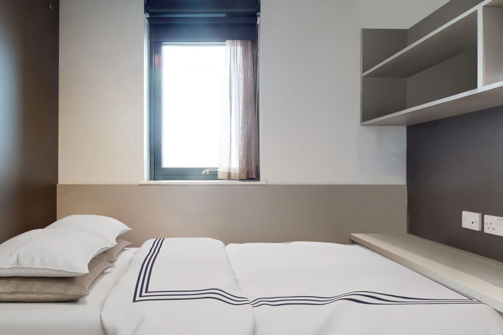Ein Hotelzimmer mit Bett, Fenster mit Vorhängen, Ablage über dem Bett und einer Steckdose.