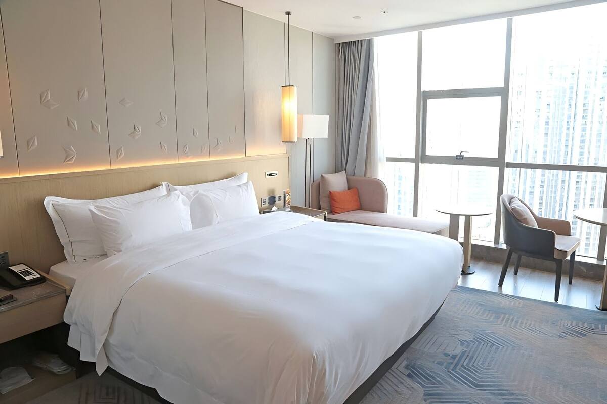 Habitación de hotel con una cama grande, un escritorio, una silla, una mesita y un ventanal con vistas a la ciudad.