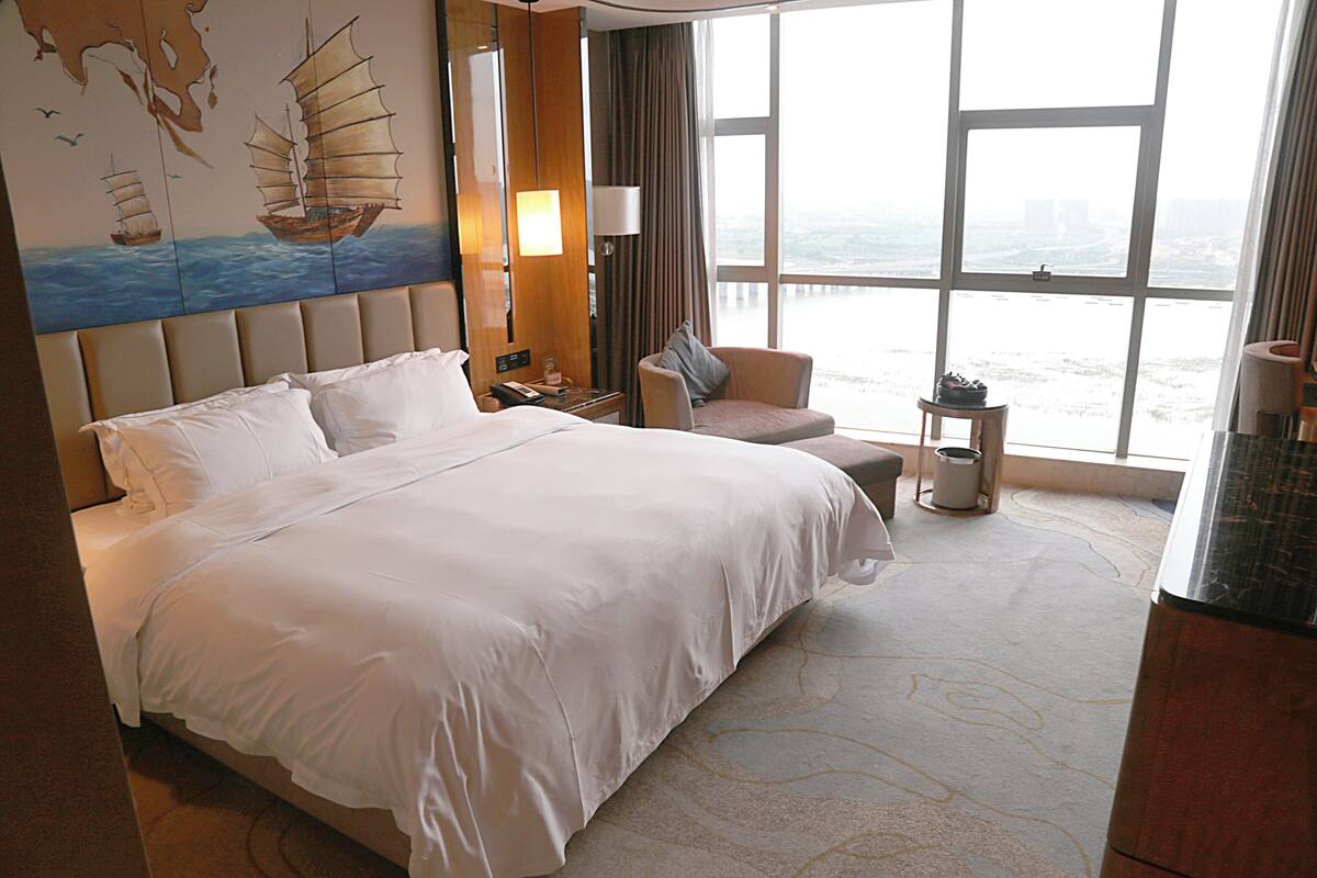 Habitación de hotel con una cama grande, una silla, una mesita y vistas de la ciudad a través de amplias ventanas.