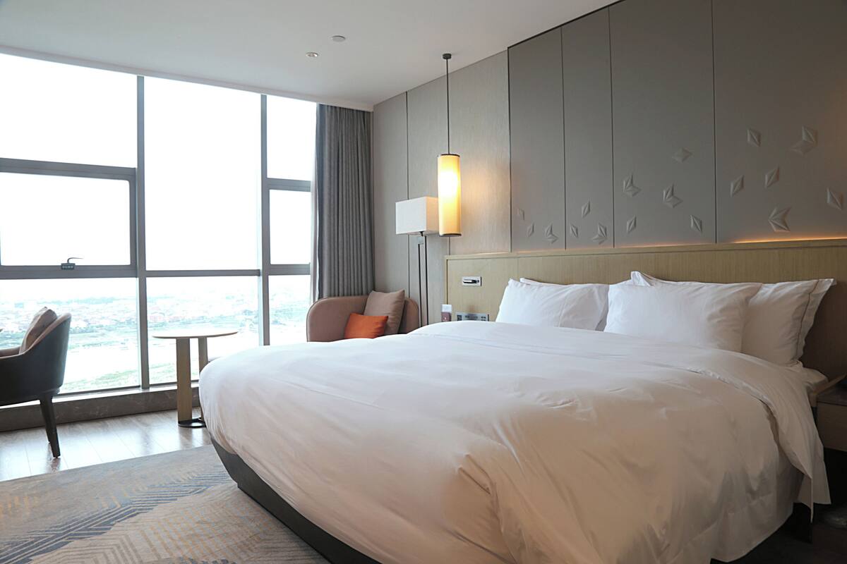Habitación de hotel con una cama grande, una silla, una mesita y vistas a la ciudad a través de amplios ventanales.