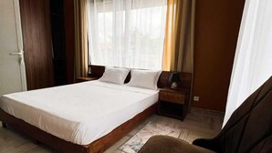 Un dormitorio con una cama grande, suelo de madera, un sillón marrón, una mesita de noche con lámpara y una ventana con cortinas.