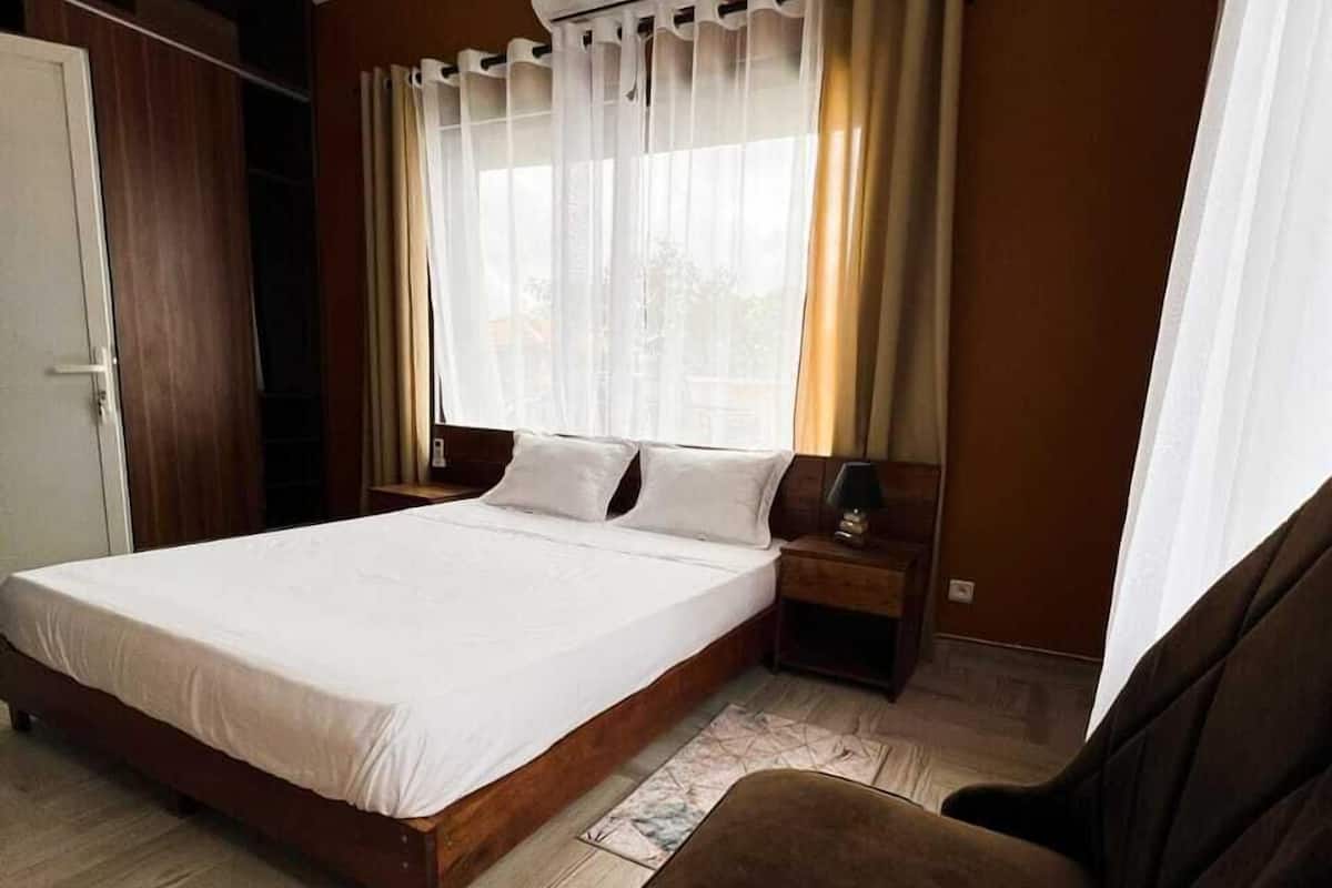 A bedroom with a large bed, a wooden floor, a brown armchair, a bedside table with a lamp, and a window with curtains.