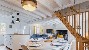A modern kitchen with white cabinets, a dining table set for four, and a wooden staircase. - Charming Cottage with Terrace, Near Beaches, Parking, Wi-Fi, Baby Equipment, and Ping-Pong (Saint-Lyphard)