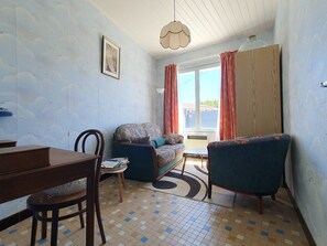 Une pièce comprenant une table à manger, deux canapés, un fauteuil, une petite table avec des livres, un sol à motifs, un luminaire au plafond et une fenêtre avec des rideaux.