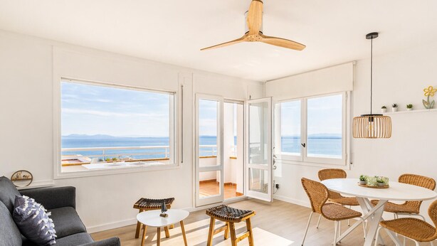 A modern living room with a sofa, a dining table with chairs, a ceiling fan, and large windows offering a view of the sea. - 2-Bedroom Apartment with Beach Access & Private Parking (Roses)