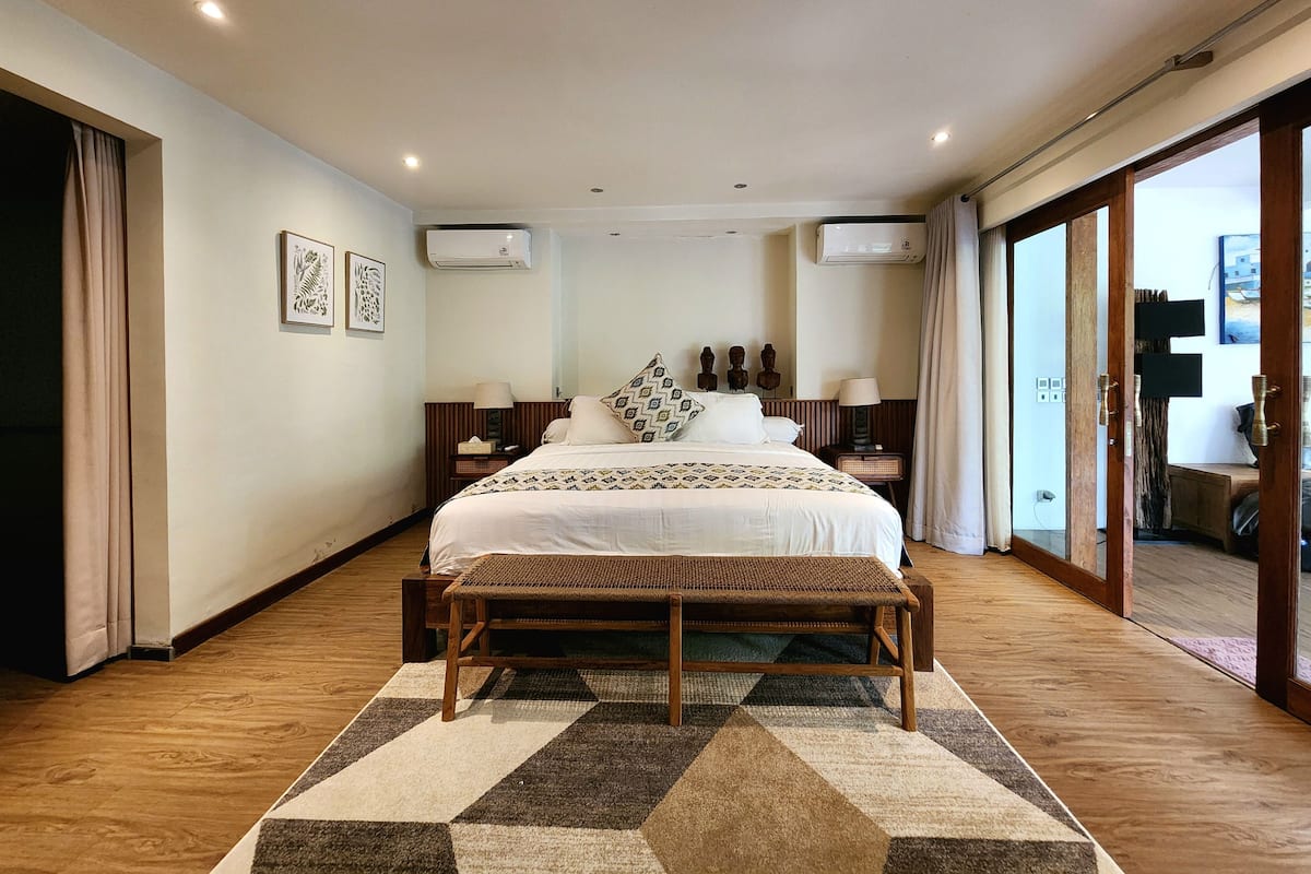 A bedroom with a large bed, a wooden bench, a patterned rug, and a view of the outside.