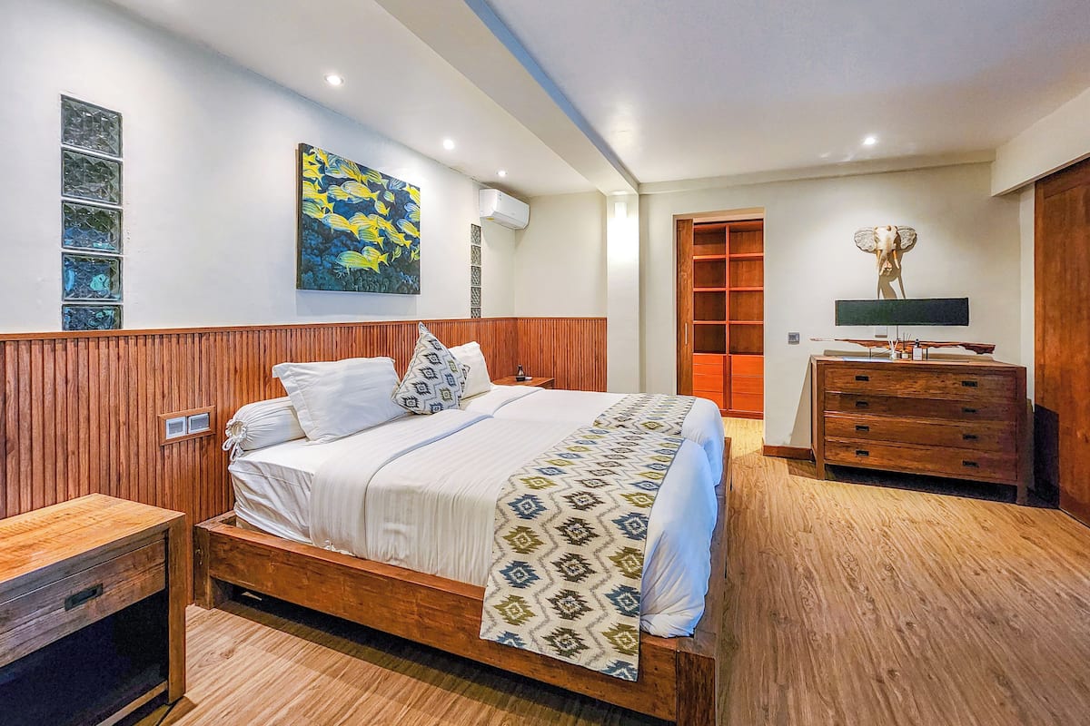 A bedroom with a wooden bed, a nightstand, a dresser, a wall-mounted TV, and a decorative sculpture.