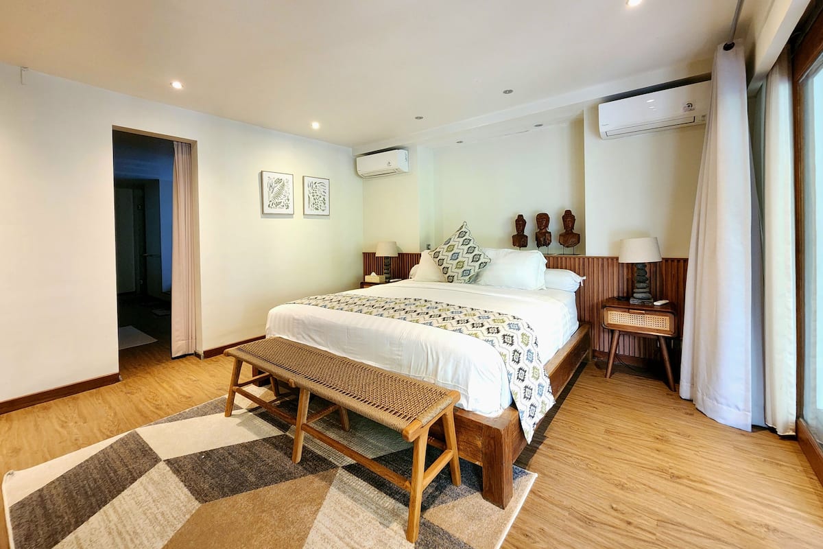 A hotel room with a large bed, a bench, a bedside table with a lamp, and a wall-mounted air conditioner.