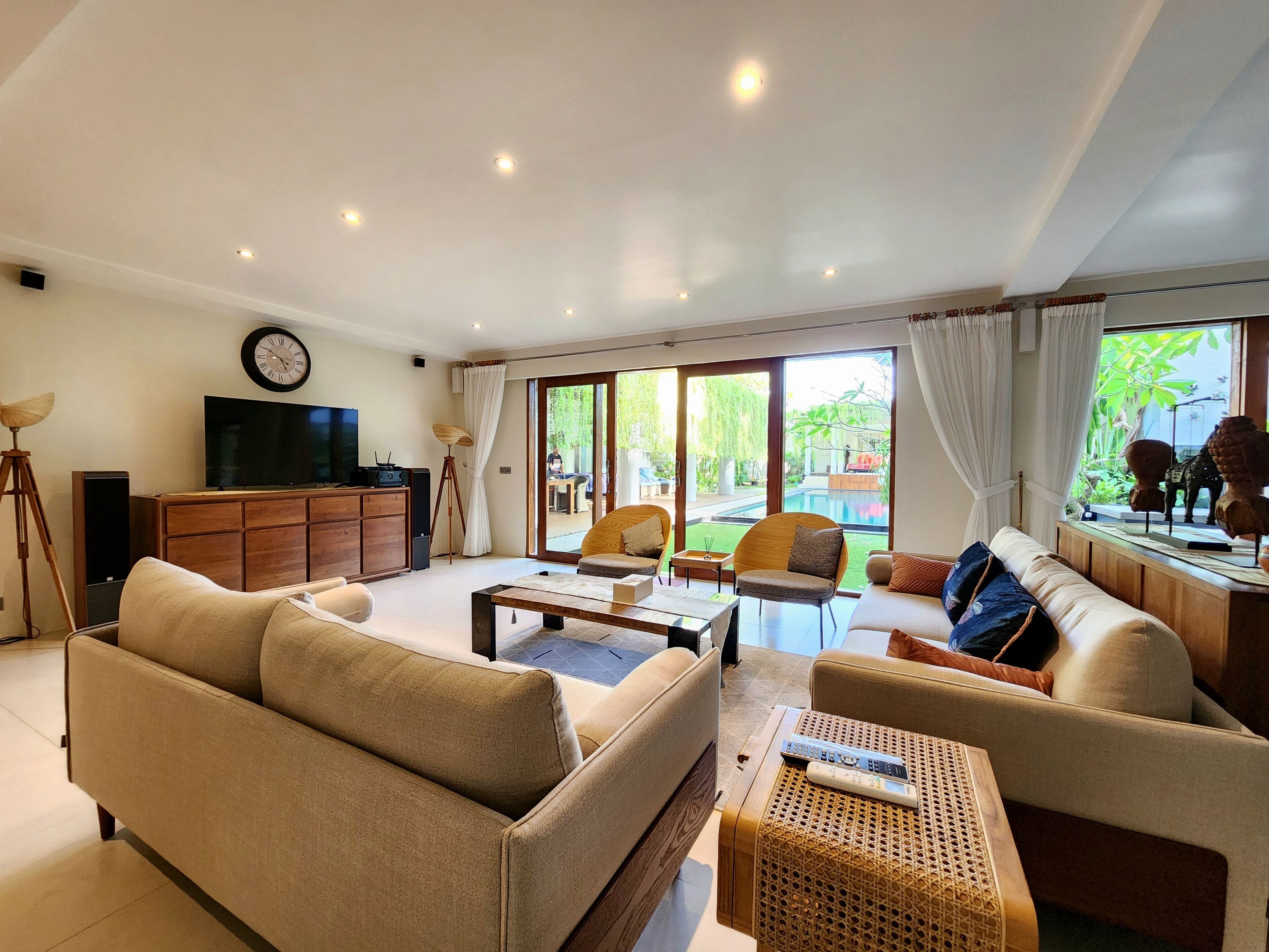 A spacious living room with a large sofa, a coffee table, and a television on a wooden cabinet. There are large windows with curtains, and a view of a pool and greenery outside.