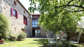 A stone building with a modern glass extension, a patio with metal chairs, and a tree providing shade.