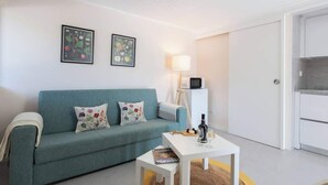 Un salon avec un canapé de couleur turquoise, deux coussins à motifs floraux, une table basse blanche sur laquelle se trouve un magazine, une lampe et un four à micro-ondes.