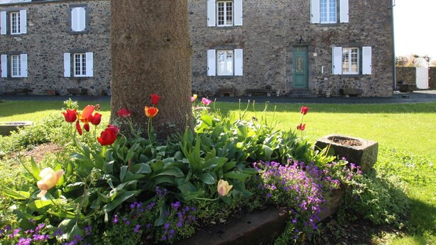 Un edificio de piedra con múltiples ventanas y contraventanas, rodeado por un jardín bien cuidado con diversas flores y un árbol.
