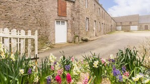 Un bâtiment en pierre avec une porte blanche et un volet en bois, entouré d’une clôture en bois peinte en blanc et de fleurs aux couleurs vives.