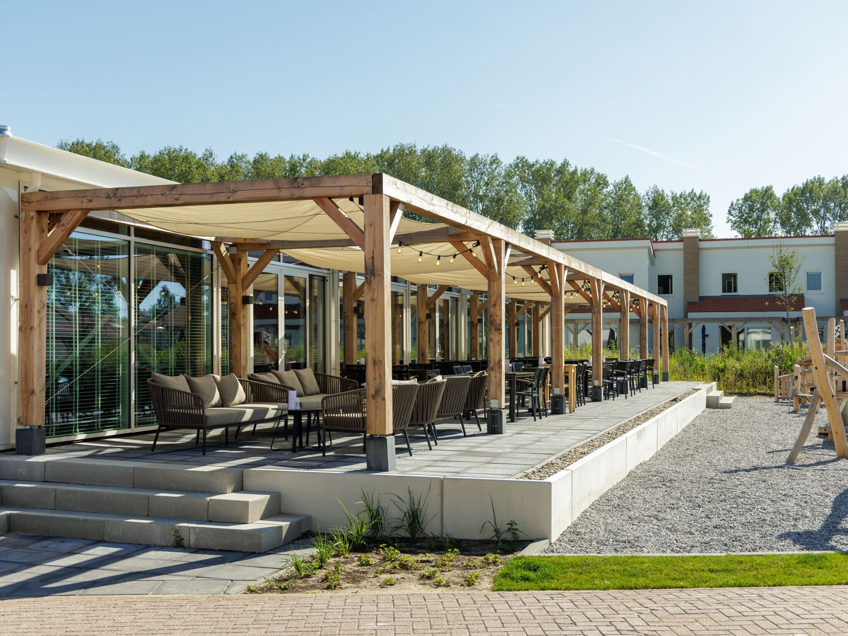 An outdoor patio area with wooden pergolas, seating arrangements, and a gravel path leading to a building.