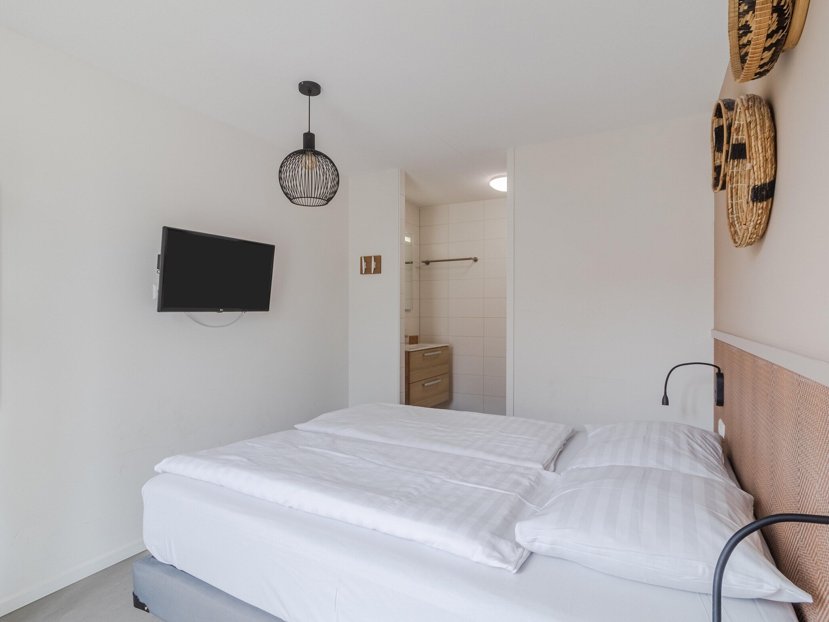 A bedroom with a bed, a television mounted on the wall, a hanging light fixture, and a bathroom visible through an open door.