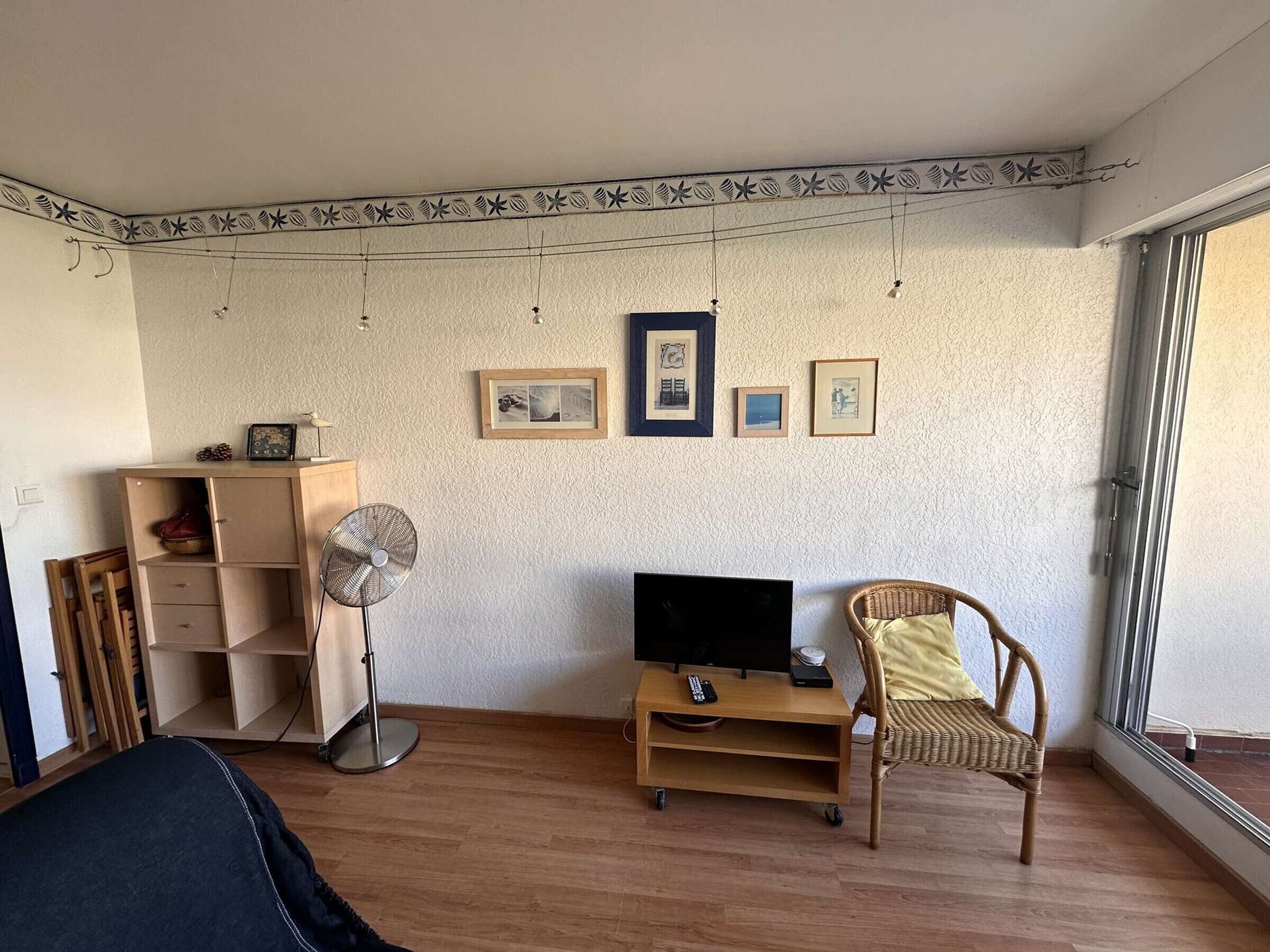 Una habitación con suelo de madera, un televisor sobre un mueble bajo, un ventilador, una silla y una ventana.
