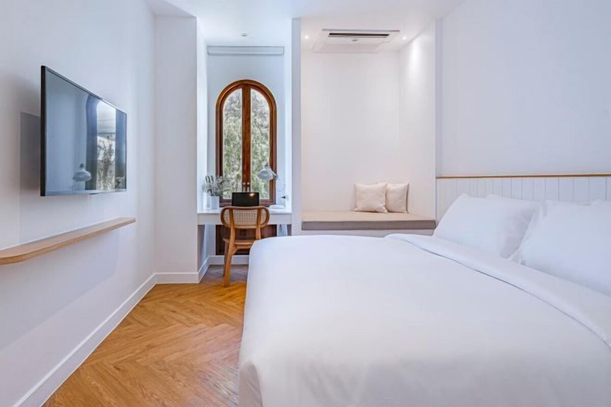 A modern hotel room with a large bed, a television mounted on the wall, a wooden floor, and a window with a view of greenery.
