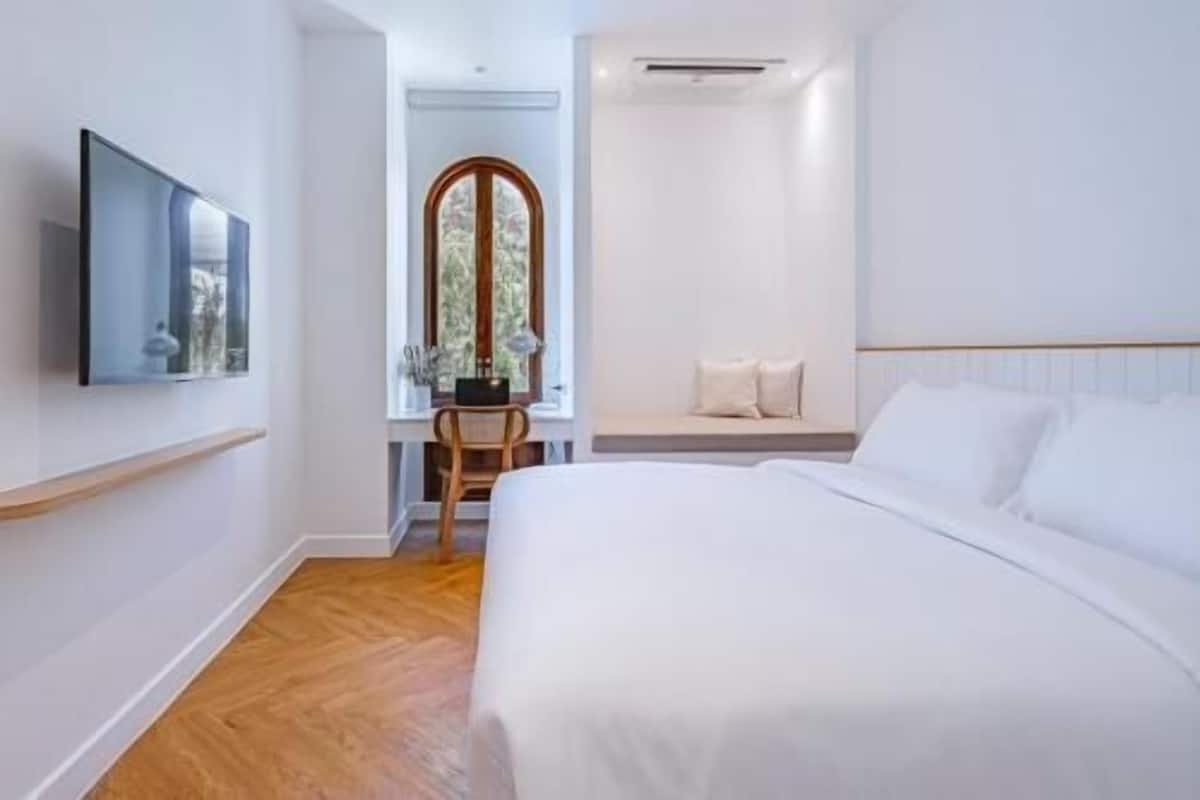 A modern hotel room with a large bed, a flat-screen TV mounted on the wall, a wooden chair, and a window with a view of greenery.