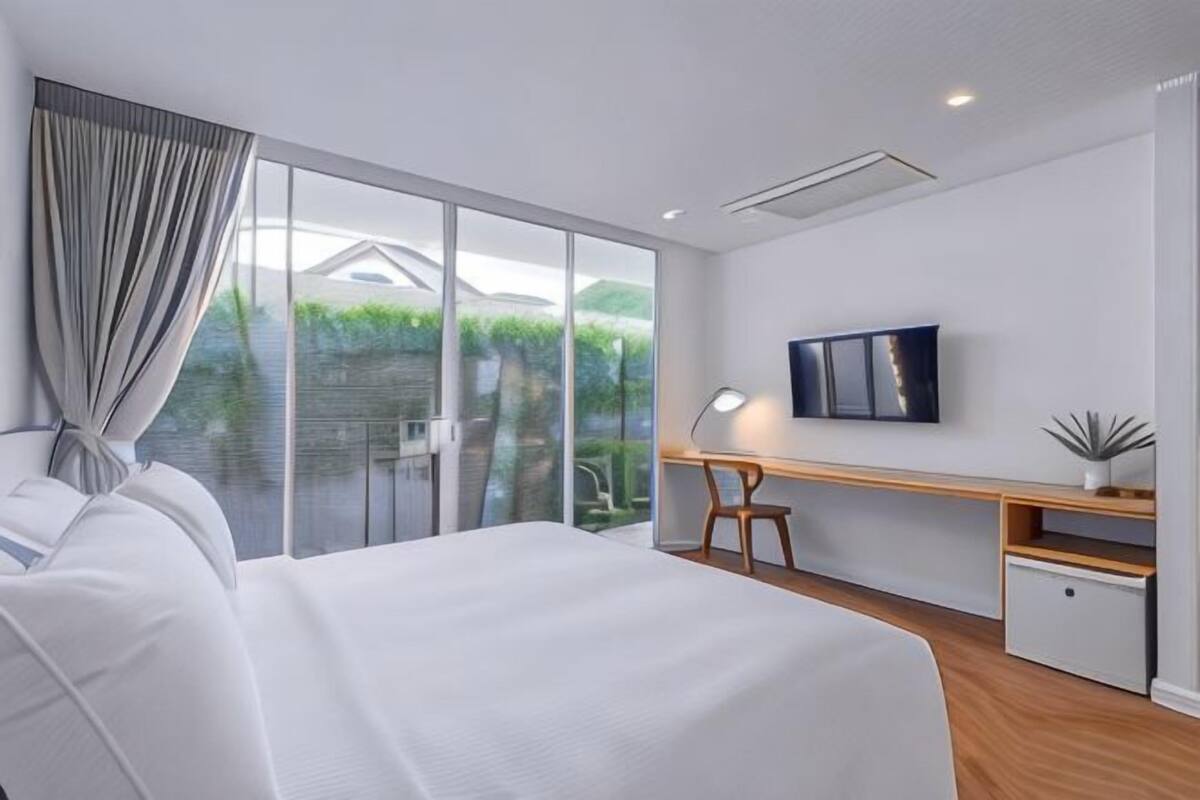 A modern hotel room with a large bed, a desk, a TV, and a sliding glass door to the outdoors.