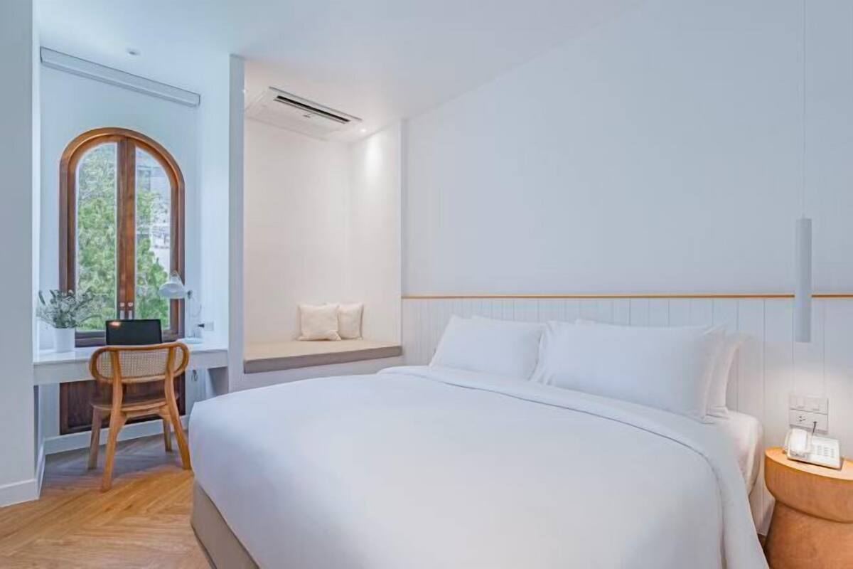 A modern bedroom with a large bed, a desk with a chair, a window, and a bedside table with a lamp.