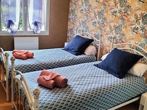 A room with two beds, each with a patterned bedspread and pillows, a window with sheer curtains, and a floral wallpaper.