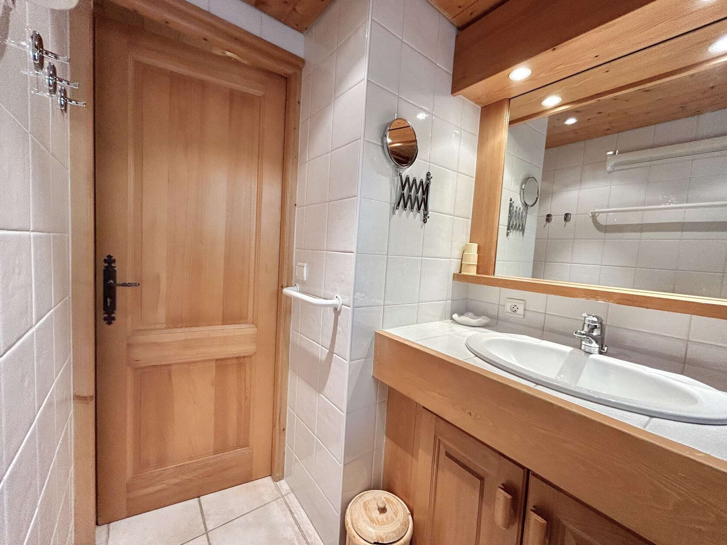 Une salle de bain avec une porte en bois, des murs recouverts de carreaux blancs, un lavabo avec un miroir et un porte-serviettes.