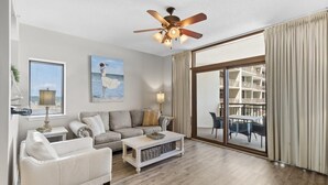 A spacious living room with a ceiling fan, a large window with curtains, a sofa, a coffee table, and a painting of a woman by the sea. - Spectacular 8th Floor Serenity: Oceanfront Views (North Myrtle Beach)