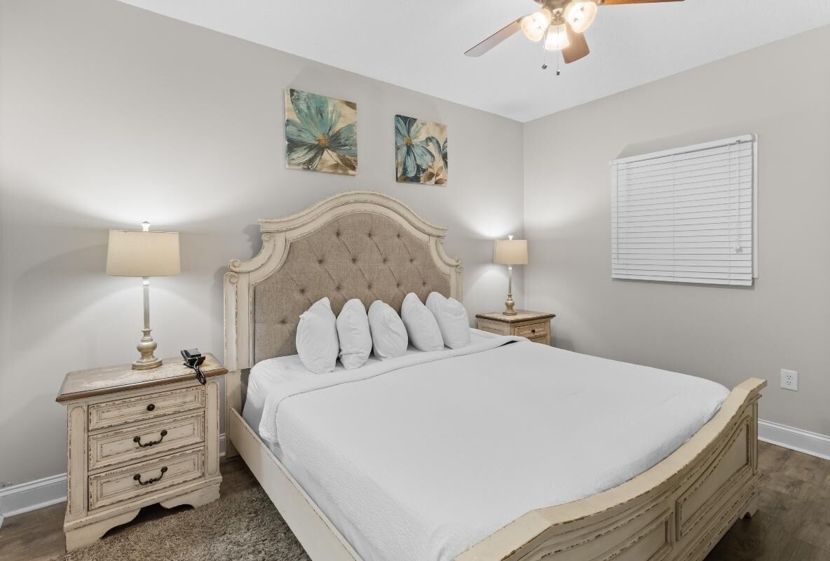 Une chambre à coucher avec un grand lit, deux tables de chevet avec des lampes, un ventilateur de plafond et deux tableaux accrochés au mur.