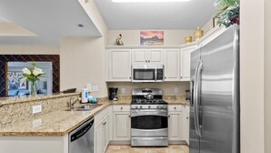 Una cocina compacta con armarios blancos, electrodomésticos de acero inoxidable, encimeras de granito y suelo de baldosas.