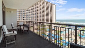 Un balcón con vista a un complejo turístico en la playa, que incluye una zona de piscina y un rascacielos al fondo.