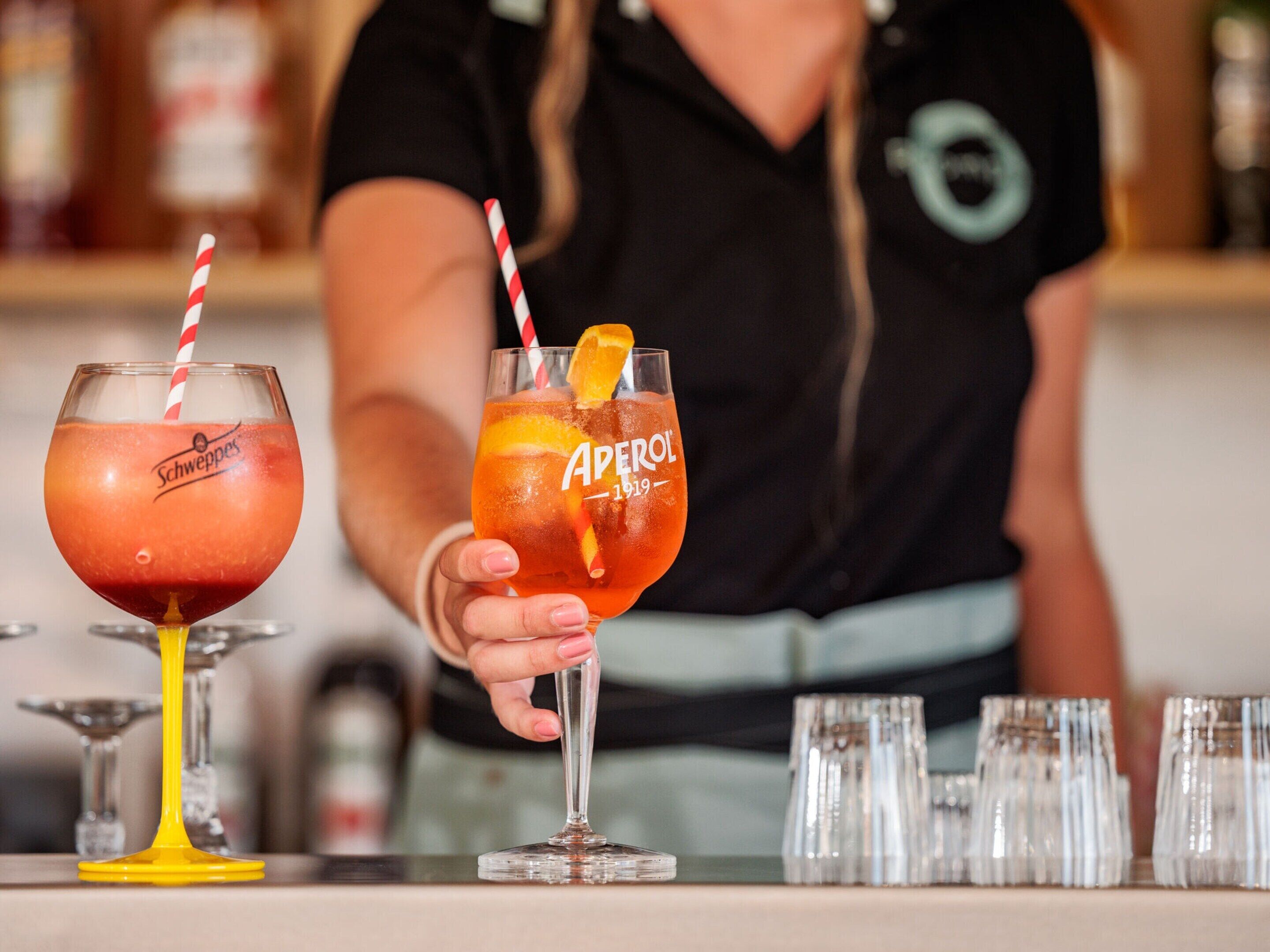 Two cocktails in glasses with brand names visible, held by a person in a black shirt with a logo.