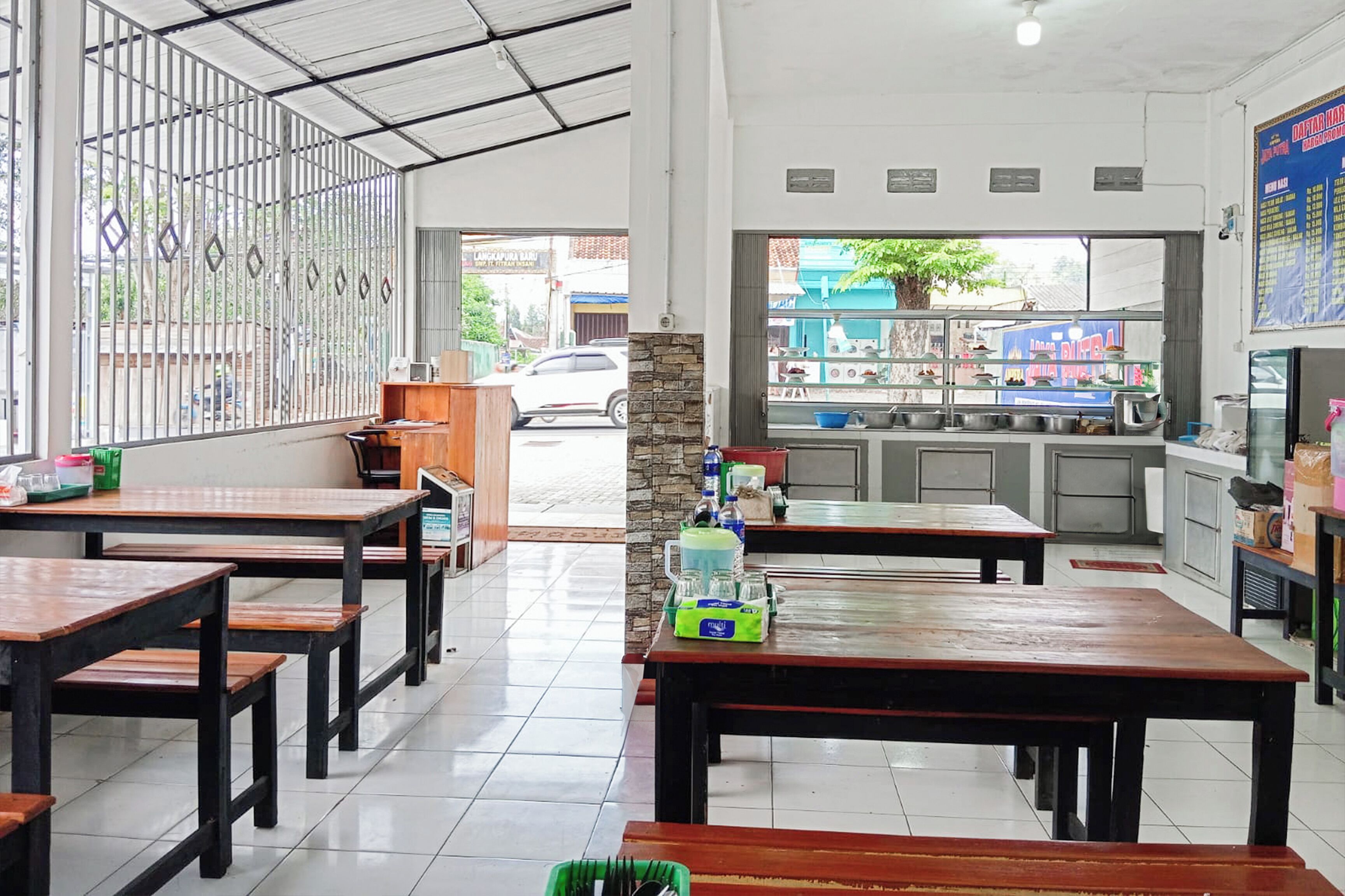 A restaurant interior with wooden tables and benches, a kitchen area visible through a window, and a metal fence outside.
