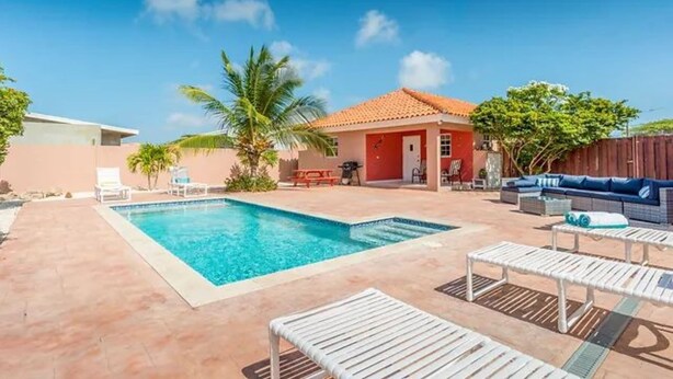 A backyard with a swimming pool, lounge chairs, and a red-roofed building. - Ideal Home for Larger Family with Pool in Noord (Noord)