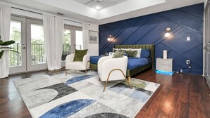 A modern bedroom with a bed, a white armchair, a blue accent wall, and a view to the outside.