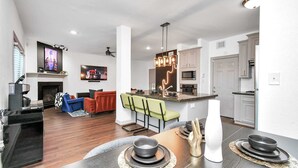 A modern living room with a fireplace, a dining area with a table set for four, and a kitchen with white cabinets and a bar area.