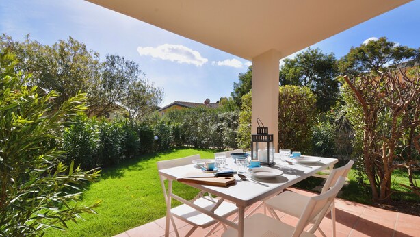 Un patio esterno con tavolo bianco apparecchiato per mangiare, immerso nel verde e con cielo sereno.