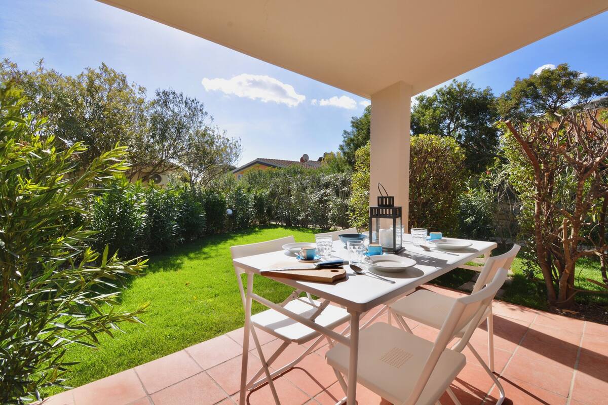 Un patio al aire libre con una mesa blanca lista para comer, rodeado de abundante vegetación y un cielo despejado.