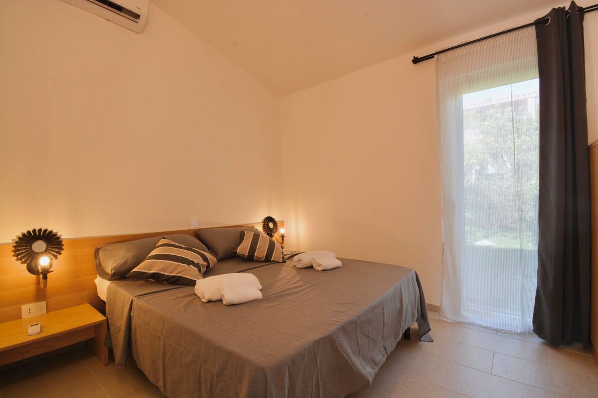 Un dormitorio con cama, mesitas de noche, aire acondicionado de pared y una ventana con cortinas.