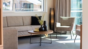 A modern living room with a sofa, a round coffee table, and a chair. There is a large window with curtains, a floor lamp, and a view of buildings outside. - Villa in Breskens Near the Beach (Breskens)