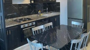 Uma cozinha moderna com armários escuros, bancada de mármore, forno embutido e um espaço de jantar com mesa de mármore preto e cadeiras brancas.