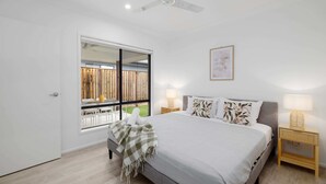 Un dormitorio con cama, lámparas de noche, ventilador de techo, ventana con vista a una valla de madera y un cuadro en la pared.