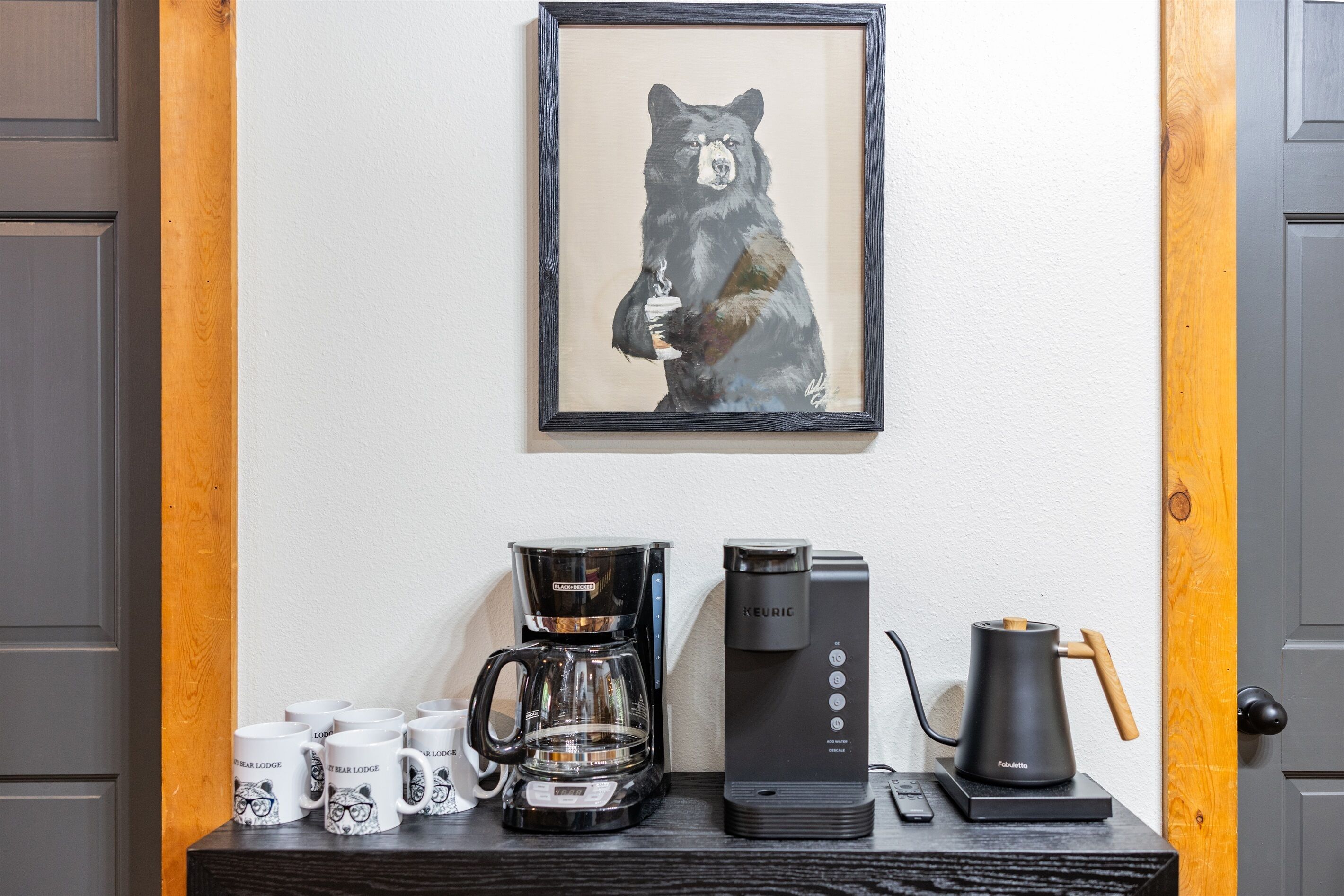 Angolo caffè con una macchina da caffè Keurig, una caffettiera a stantuffo e diverse tazze su un tavolo di legno, davanti a un muro bianco con un quadro di un orso incorniciato.