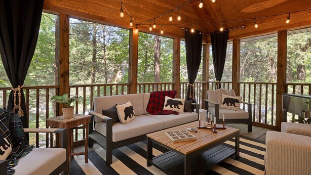 Una terraza de madera con sofá, sillones y una mesa, rodeada de amplios ventanales con vistas a árboles.