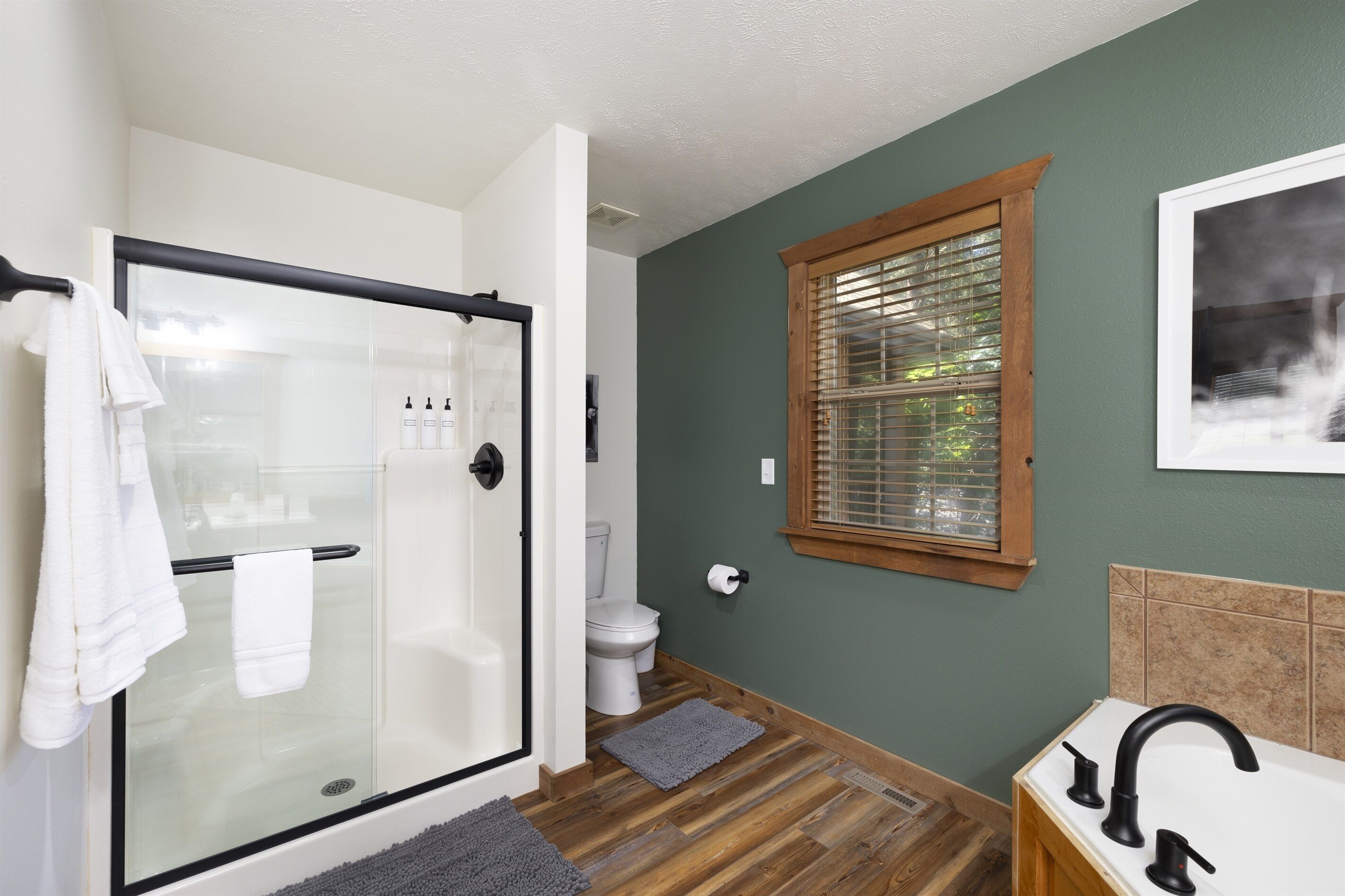 A bathroom with a glass shower enclosure, a white toilet, a wooden vanity with a black faucet, and a window with blinds.